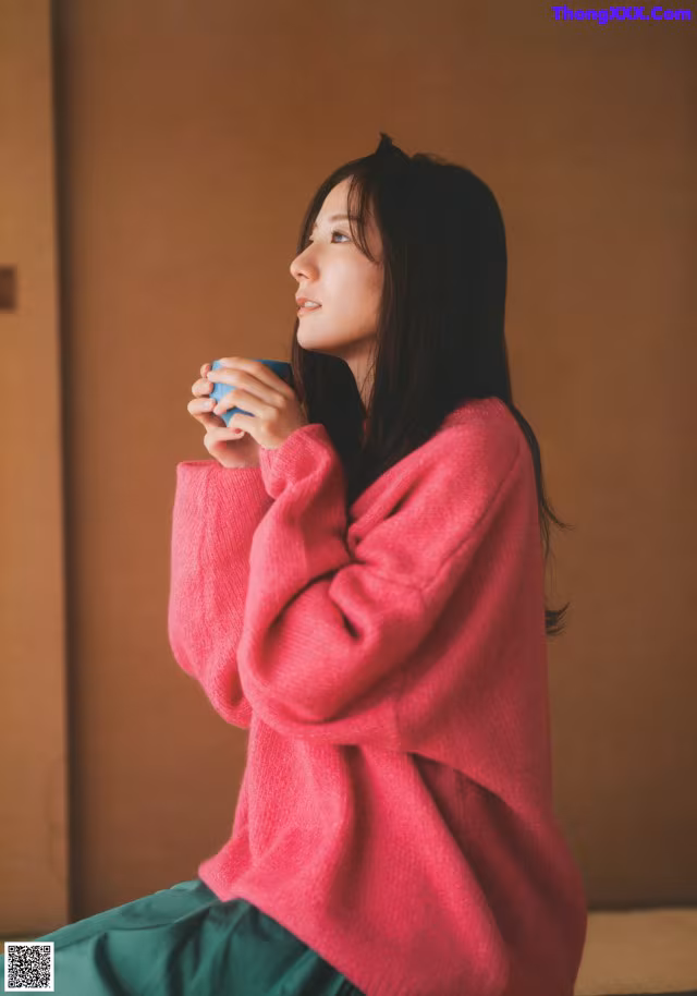 A woman in a pink sweater sitting on a bed holding a cup of coffee.
