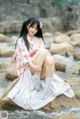 A woman in a kimono sitting on a rock by the water.